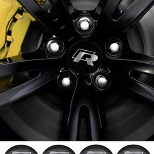 Könnte beinhalten: Ein Set aus vier runden, schwarzen Radnabendeckeln mit einem silbernen R-Logo. Die Deckel werden unter einer Nahaufnahme eines schwarzen Fünfspeichen-Autofelgenrads mit passender Nabenkappe und einem gelben Bremssattel gezeigt.