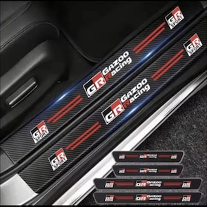 May include: A set of black carbon fiber textured door sill protectors for a vehicle. Each piece features the red and white GR Sport logo, the Gazoo Racing text, and two parallel red stripes. The design is sleek, durable, and automotive-focused.
