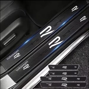 Puede incluir: Un juego de cuatro protectores de umbral de puerta con textura de fibra de carbono negra para vehículo. Cada tira rectangular presenta el logotipo blanco de Volkswagen y la letra R impresos en la superficie. Los protectores están instalados.