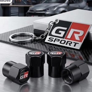May include: A metal keyring featuring a rectangular plate with the text GR SPORT in white and red against a black background, accompanied by four black hexagonal tyre valve caps, each displaying a small GR logo on the top surface.
