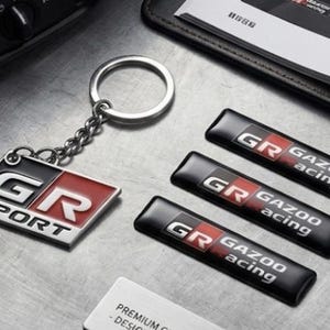May include: A metal keyring featuring a rectangular GR SPORT emblem with red, white, and black details, accompanied by three rectangular black adhesive badges displaying the GR GAZOO RACING logo in white and red lettering on a metallic surface.