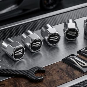 Könnte beinhalten: Fünf metallisch silberne Ventilkappen mit schwarzer runder Einlage und weißem, stilisiertem R-Logo, dazu ein Schraubenschlüssel in Carbonfaser-Optik und zwei rechteckige schwarze Embleme mit weißem R-Logo auf einer Metall- und Holzoberfläche.