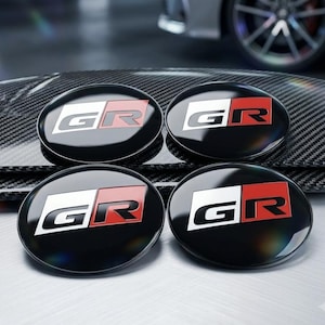 May include: Four circular automotive emblems featuring the GR logo in black, white, and red. Each emblem has a glossy black finish and displays the letters G and R in a bold, stylized font. The emblems are arranged in a two-by-two grid pattern.