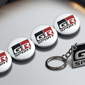 May include: Three circular metallic emblems and one rectangular metal keyring, each featuring the GR SPORT logo in black, white, and red. The circular emblems have a polished silver finish, while the rectangular keyring includes a silver ring.