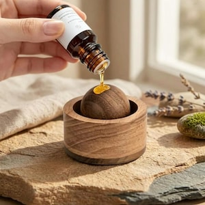 May include: A small amber glass bottle with a white label is held above a wooden diffuser set. The set consists of a circular wooden base containing a smooth, dark brown wooden sphere. A single drop of golden essential oil falls from the bottle.