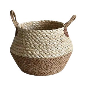 May include: A round woven basket featuring a two-tone design with a light beige upper section and a darker brown lower section. The basket includes two sturdy handles attached with brown leather-like tabs on opposite sides of the rim.