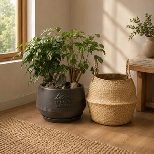 May include: A dark gray, round ceramic planter with a saucer and the embossed word FLOWER on the front, containing a green leafy plant. Beside it sits a tall, woven, light tan seagrass basket with two upright handles and a tapered middle section.