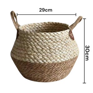 May include: A round woven basket featuring a two-tone design with a light beige upper section and a darker brown lower section. The basket includes two side handles with brown leather-like accents. Dimensions are 29 cm in width and 30 cm in height.