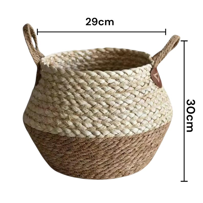 May include: A round woven basket featuring a two-tone design with a light beige upper section and a darker brown lower section. The basket includes two side handles with brown leather-like accents. Dimensions are 29 cm in width and 30 cm in height.