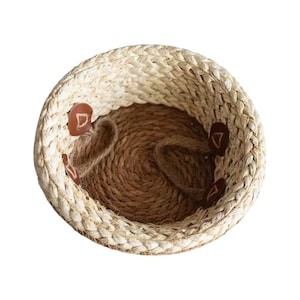 May include: A round woven basket featuring a light beige braided rim and a dark brown coiled base. The interior walls are accented with four small, circular brown leather-like patches, each embossed with a simple triangular geometric symbol.