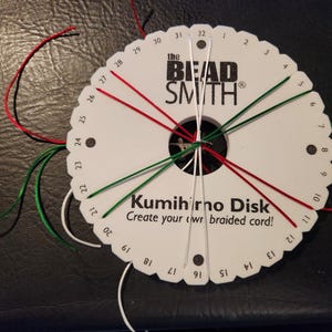 May include: A white circular Kumihimo disk by The Bead Smith with numbered slots from 1 to 32. Red, green, and white cords are threaded through the slots and meet in the centre hole. The text reads The Bead Smith Kumihimo Disk Create your own braided cord.