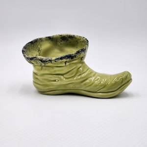 May include: A ceramic boot-shaped planter in a light olive green glaze. The top rim features a distressed black speckled finish, while the body of the boot displays molded horizontal folds and creases, giving it a textured, worn appearance.