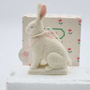 May include: A white, hand-painted bisque porcelain Easter bunny figurine with pink detailing. The bunny has textured fur, a pink ribbon, and small wings. It sits on a grassy base, with a tag and a box in the background.
