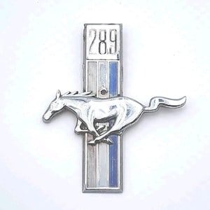 May include: A chrome Mustang emblem featuring a running horse. The emblem has a vertical bar with the number "289" at the top, and stripes of silver, white, and blue. This car badge is designed for a vehicle.
