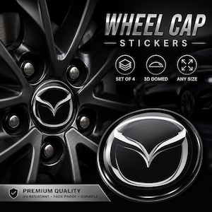 Op de afbeelding: Wieldopstickers met het Mazda-logo. De afbeelding toont een close-up van een autowiel met een zilver en zwart ontwerp. De tekst luidt "Wheel Cap Stickers", "Set of 4", "3D Domed" en "Any Size". De stickers zijn van premium kwaliteit, UV-bestendig, kleurecht en duurzaam.