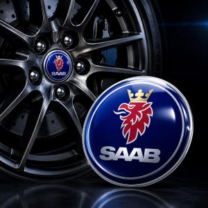 May include: A close-up of a car wheel with a silver finish and a blue and silver Saab logo. The logo features a red griffin with a gold crown and the word "SAAB" in silver lettering. The background is black.