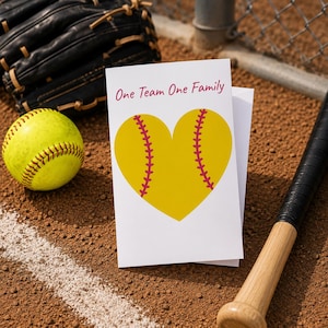 May include: A softball-themed greeting card with the text "One Team One Family" in pink. The card features a yellow heart with pink stitching, resembling a softball. A yellow softball, a black baseball glove, and a baseball bat are also visible.