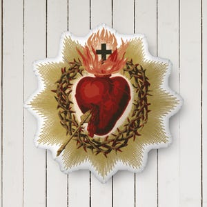 May include: A religious emblem featuring a red heart pierced by an arrow, encircled by a crown of thorns, and topped with a cross and flames. The heart is set against a gold burst design on a white background.