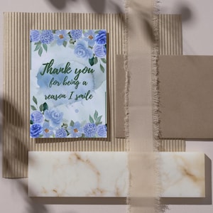 Op de afbeelding: Een wenskaart met blauwe bloemaccenten en de tekst "Thank you for being a reason I smile". De kaart is omgeven door een beige envelop, een marmerachtig blok en een textuurachtergrond.