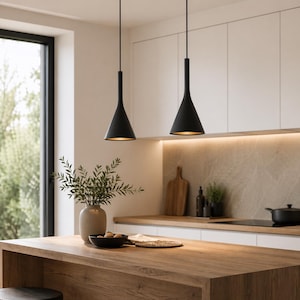 Può includere: Due lampade a sospensione nere con paralume a forma di cono sono appese sopra un'isola cucina in legno. Le luci hanno un design minimalista, con un gambo stretto e una base più ampia. La cucina presenta mobili bianchi, un paraschizzi in marmo e un vaso con vegetazione.