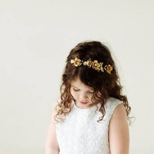 May include: A young girl wearing a white dress with a gold floral crown in her hair. She is looking down at her hands.