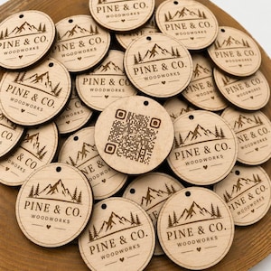 May include: A collection of round wooden tags with the text "PINE & CO. WOODWORKS" and a mountain and tree design. One tag features a QR code. The tags are arranged in a wooden bowl.