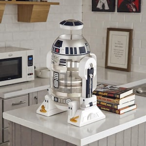 Peut inclure: Une cafetière à piston R2-D2 argentée et blanche. La cafetière a un corps cylindrique avec des accents bleus et noirs, une base blanche et une poignée. C'est un appareil de cuisine.