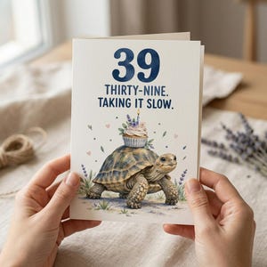 Peut inclure: Une carte de vœux blanche avec le grand nombre 39 en bleu foncé et le texte THIRTY-NINE. TAKING IT SLOW. Sous le texte, une illustration montre une tortue portant un cupcake avec une bougie allumée sur sa carapace, entourée de brins de lavande.