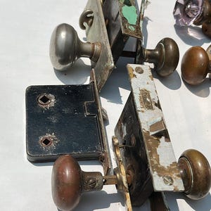 Op de afbeelding: Een verzameling vintage metalen insteeksloten en diverse deurknoppen in messing, stalen en glazen afwerkingen. Het hang- en sluitwerk bevat rechthoekige voorplaten, interne slotmechanismen en ronde knoppen met tekenen van slijtage en patina.