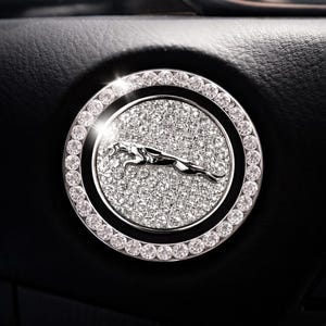 May include: A circular car emblem featuring a silver Jaguar logo. The emblem is surrounded by a ring of clear, sparkling crystals and a background of smaller, glittering crystals. The emblem is set against a black background.