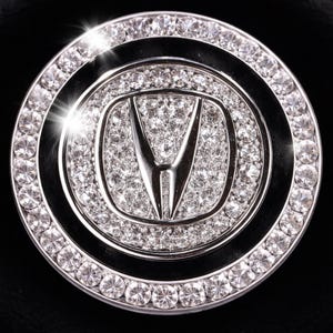 May include: A round, silver-toned emblem with a central logo and a sparkling, crystal-studded border. The emblem features a silver logo in the center, surrounded by a ring of clear crystals. The outer edge is also lined with crystals, creating a dazzling effect.