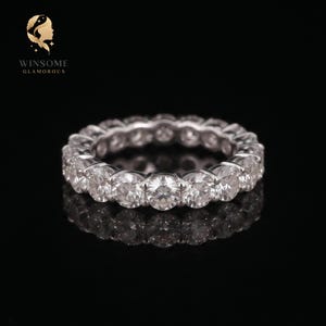 May include: A silver-toned eternity band featuring a continuous row of round, clear, faceted stones set in a polished metal frame. The ring is displayed against a black background with the text WINSOME GLAMOROUS visible in the upper left corner.