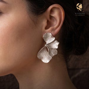 May include: A silver-toned earring featuring a layered, fan-like design with delicate, etched vertical lines resembling organic leaf structures. The piece has a polished metallic finish and is secured to the earlobe with a post-style backing.