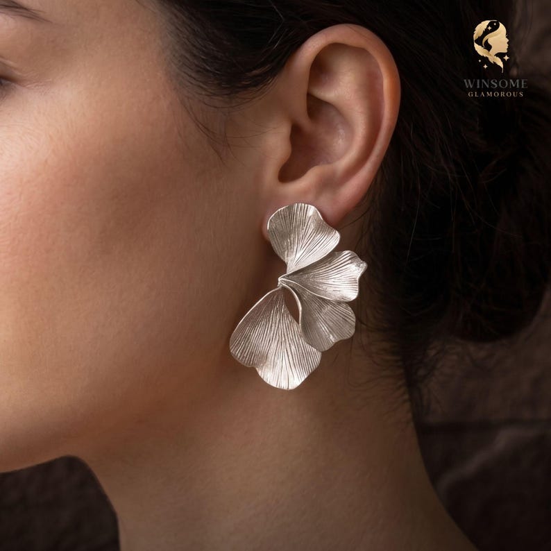 May include: A silver-toned earring featuring a layered, fan-like design with delicate, etched vertical lines resembling organic leaf structures. The piece has a polished metallic finish and is secured to the earlobe with a post-style backing.