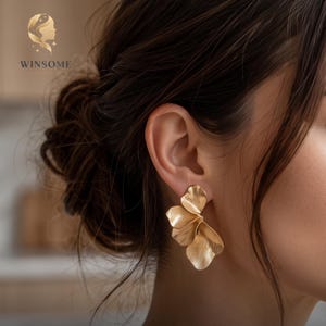 May include: A gold-toned earring shaped like a flower with multiple overlapping petals. The surface of the metal features fine, linear texturing that radiates from the center of each petal, creating a layered, fan-like appearance on the earlobe.