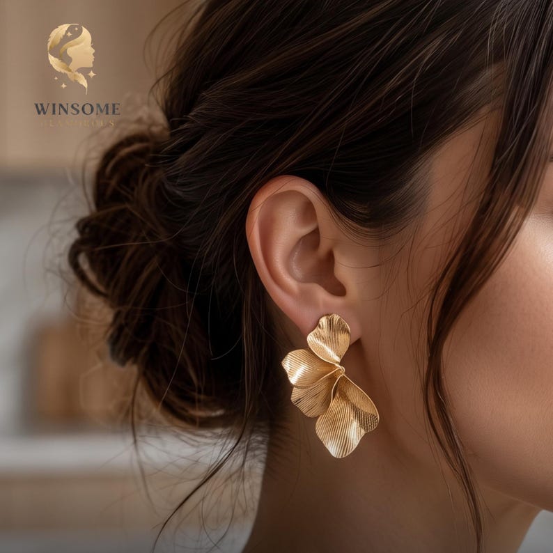 May include: A gold-toned earring shaped like a flower with multiple overlapping petals. The surface of the metal features fine, linear texturing that radiates from the center of each petal, creating a layered, fan-like appearance on the earlobe.