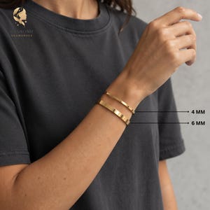May include: Two gold-toned metal cuff bracelets worn on the wrist. The top bracelet has a width of 4 mm, and the bottom bracelet has a width of 6 mm. Both bracelets feature a smooth, polished finish and a simple, open-ended design.