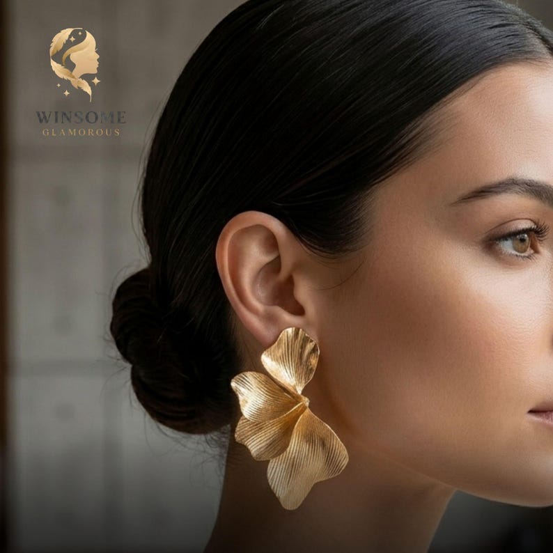 May include: A gold-toned earring shaped like a large, textured flower petal. The piece features a brushed metallic finish with delicate, radiating lines that mimic the organic veins of a leaf or petal. The logo WINSOME GLAMOROUS is visible.