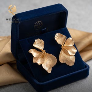 May include: A pair of gold-toned earrings shaped like layered flower petals with a textured, ribbed surface. The earrings are displayed inside a dark blue velvet jewelry box that features the text WINSOME GLAMOROUS and a logo.