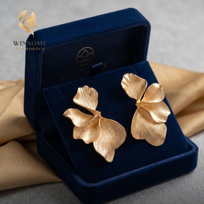 May include: A pair of gold-toned earrings shaped like layered flower petals with a textured, ribbed surface. The earrings are displayed inside a dark blue velvet jewelry box that features the text WINSOME GLAMOROUS and a logo.