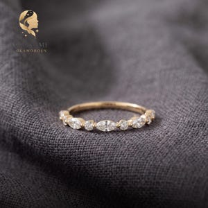May include: A gold band ring featuring an alternating pattern of marquise-cut and round-cut clear stones set along the top half of the band. The ring is positioned horizontally, showcasing the clear, faceted stones and the polished gold metal finish.
