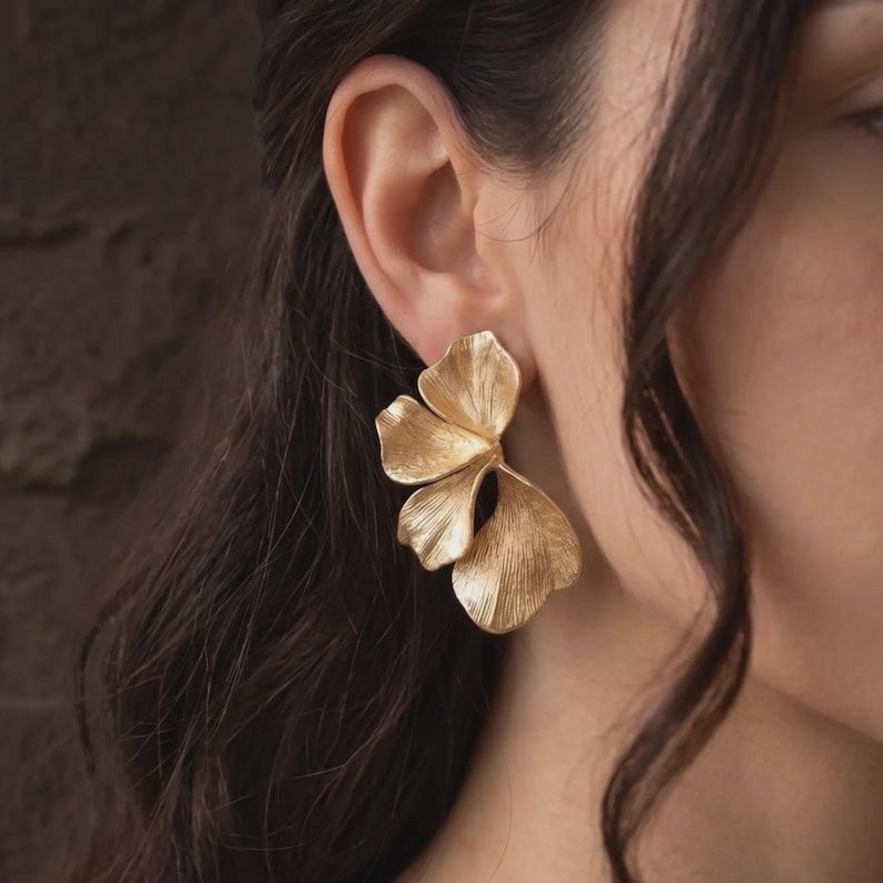 May include: A gold-toned earring featuring a layered, fan-like design with three distinct petal-shaped segments. The surface of the metal has a fine, linear texture that radiates from the center of each segment, creating a metallic floral appearance.