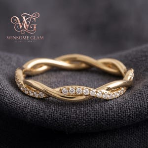May include: A gold ring featuring a twisted band design with one strand set with small, clear, round-cut stones. The ring is positioned against a dark, textured fabric background. The logo WINSOME GLAM is visible in the upper left corner.