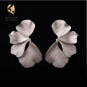 May include: A pair of silver-toned earrings shaped like ginkgo leaves with a textured, fan-like surface. The earrings feature multiple overlapping petal segments with fine, radiating lines. The logo WINSOME GLAMOROUS is visible in the top left corner.
