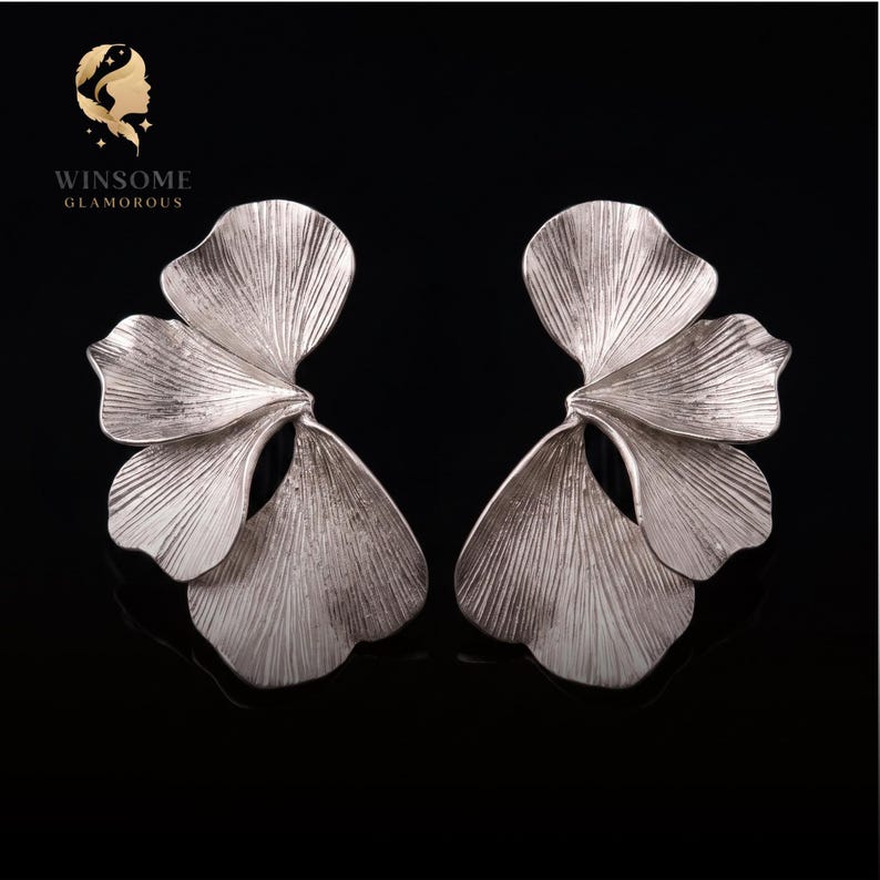 May include: A pair of silver-toned earrings shaped like ginkgo leaves with a textured, fan-like surface. The earrings feature multiple overlapping petal segments with fine, radiating lines. The logo WINSOME GLAMOROUS is visible in the top left corner.