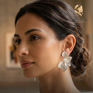 May include: A silver-toned earring shaped like a delicate, multi-layered flower petal with a matte finish. The earring features organic, flowing curves that mimic the appearance of a natural leaf or blossom, attached to the earlobe.