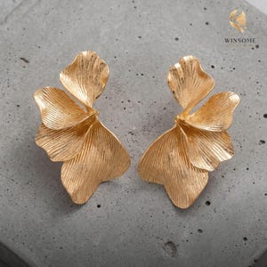 May include: A pair of gold-toned earrings featuring a layered, petal-like design with a textured, ribbed surface. Each earring consists of multiple overlapping, fan-shaped segments that create a dimensional, organic form with a metallic finish.