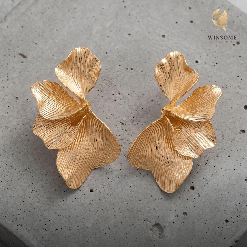 May include: A pair of gold-toned earrings featuring a layered, petal-like design with a textured, ribbed surface. Each earring consists of multiple overlapping, fan-shaped segments that create a dimensional, organic form with a metallic finish.