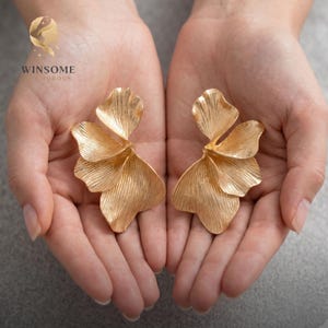 May include: A pair of gold-toned earrings shaped like layered flower petals with a textured, ribbed surface. The earrings feature a metallic finish and organic, curved edges that mimic the appearance of delicate, blooming floral petals.
