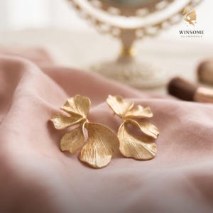 May include: A pair of gold-toned earrings shaped like delicate, layered flower petals with a textured, brushed metal finish. The earrings feature organic, curved forms that mimic the appearance of natural foliage, resting on a soft, light pink fabric.